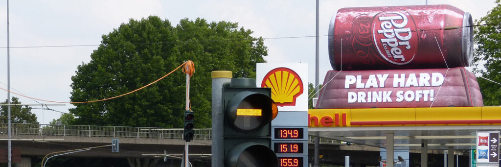 Eine Shell-Tankstelle mit großer Dr-Pepper-Werbung auf dem Dach. Das Motiv zeigt eine überdimensionierte Dose mit dem Slogan „Play Hard – Drink Soft!“. Im Vordergrund ist eine Ampel mit leuchtendem Gelb zu sehen.