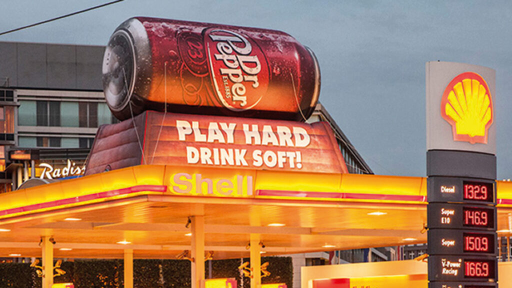 Eine Shell-Tankstelle bei Abenddämmerung, hell erleuchtet im typischen Gelb-Rot. Auf dem Dach prangt eine übergroße Dr-Pepper-Dose mit dem Slogan „Play Hard – Drink Soft!“. Daneben ist die Preistafel mit den aktuellen Kraftstoffpreisen zu sehen.
