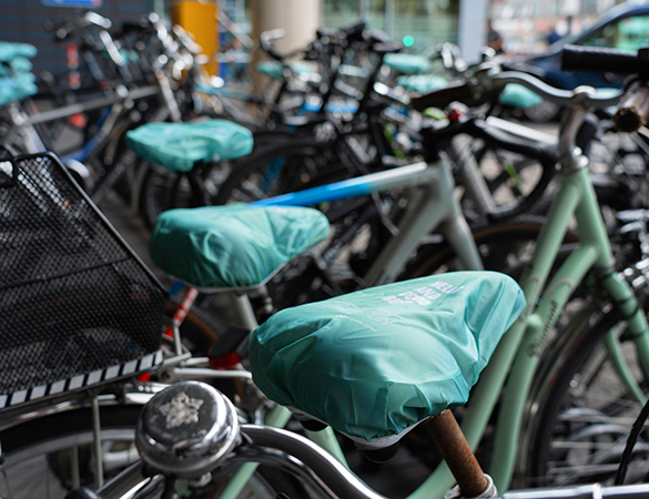 Das Bild zeigt eine Reihe von Fahrrädern, deren Sättel mit türkisfarbenen Sattelüberzügen versehen sind. Auf den Überzügen ist weiße Schrift zu sehen, die Werbung für ein Unternehmen oder eine Marke macht. Die Fahrräder stehen dicht an dicht in einem überdachten Bereich, vermutlich an einem öffentlichen Abstellplatz.