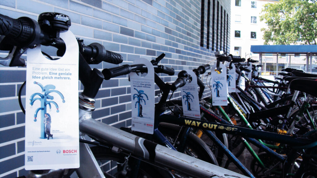 Das Bild zeigt eine Reihe von Fahrrädern, die dicht nebeneinander abgestellt sind. An vielen Lenkern hängen Werbehänger im Format von Türanhängern, die eine Bosch-Werbung tragen. Darauf steht der Slogan: „Eine gute Idee löst ein Problem. Eine geniale Idee gleich mehrere.“ Zu sehen ist außerdem ein grafisches Motiv in Blau sowie das Bosch-Logo unten rechts. Die Fahrräder stehen an einer Mauer mit blauen Kacheln.