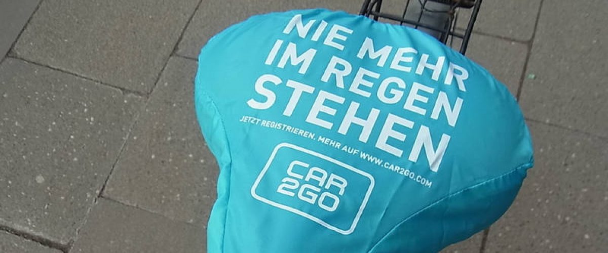 Ein türkisfarbener Fahrradsattelüberzug trägt den weißen Aufdruck „NIE MEHR IM REGEN STEHEN“ sowie das Logo von Car2Go. Darunter ist eine Webadresse mit Registrierungsaufruf angegeben.