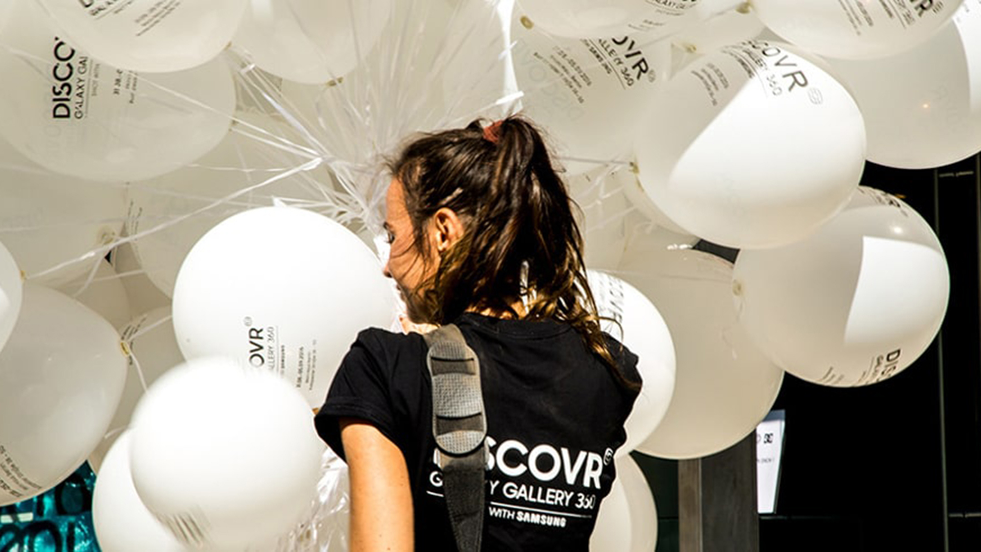 Eine junge Frau steht vor einem Bündel weißer Werbeballons mit Logoaufdruck. Sie trägt ein schwarzes Promotion-Shirt und wird von den Ballons fast vollständig umrahmt. Die Szene vermittelt Event-Charakter und Markenpräsenz.