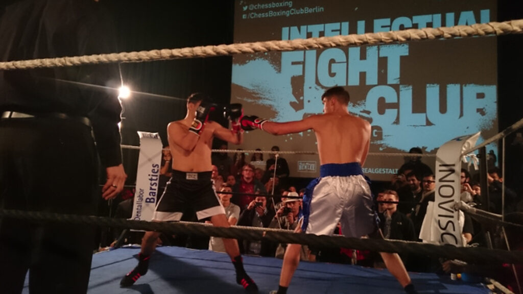 Ein Boxkampf im Ring mit großem Publikum im Hintergrund. Zwei Kämpfer stehen sich gegenüber, während auf der Leinwand der Schriftzug „Intellectual Fight Club“ erscheint. Am Ringrand ist das Logo von INOVISCO zu sehen.