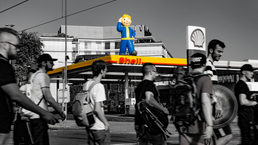 Eine Shell-Tankstelle mit farbig hervorgehobenen Gelb-Rot-Elementen im ansonsten schwarz-weißen Bild. Auf dem Dach steht eine große Figur des „Vault Boy“ aus der Fallout-Spielreihe. Im Vordergrund gehen mehrere Menschen über die Straße.