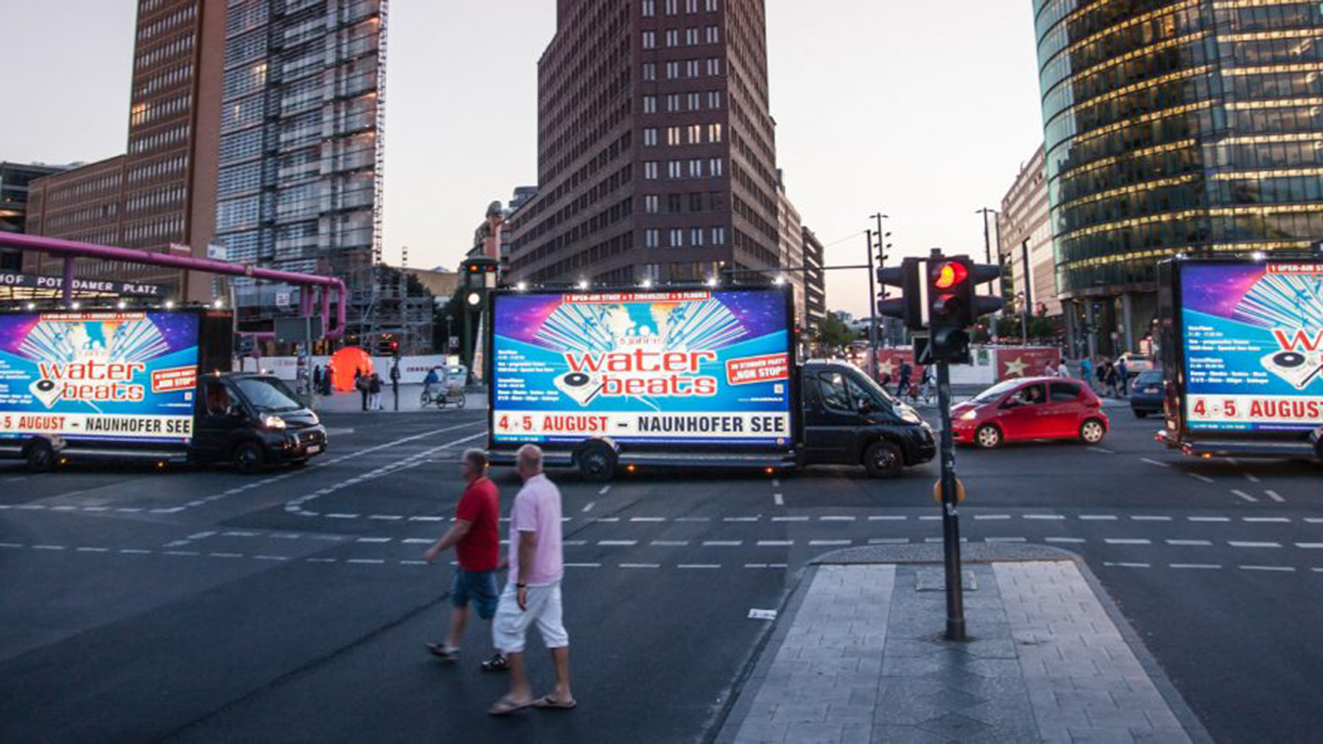 Mehrere CoolLiteTrucks von INOVISCO stehen an einer großen Kreuzung und zeigen bunte Werbeflächen für das Festival „Water Beats“ am Naunhofer See. Im Hintergrund ragen moderne Hochhäuser am Potsdamer Platz in den Abendhimmel. Fußgänger überqueren die Straße, während Autos an der Ampel warten.