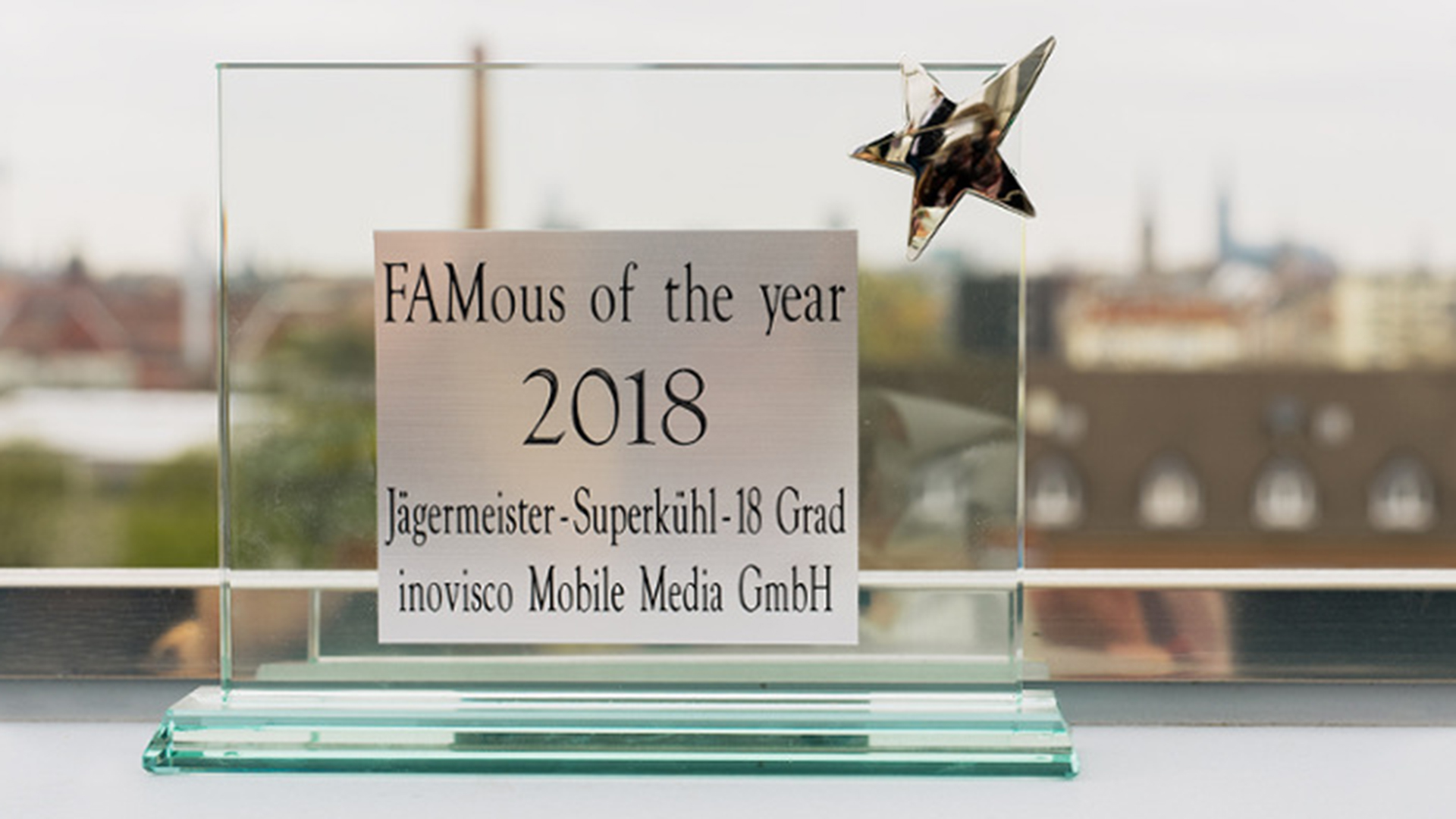 Eine gläserne Trophäe mit silberner Sternverzierung. Auf der Plakette steht „FAMous of the year 2018 – Jägermeister-Superkühl-18 Grad – inovisco Mobile Media GmbH“. Im Hintergrund ist eine verschwommene Stadtansicht zu erkennen.