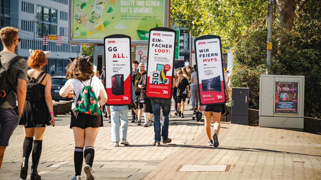 Das Bild zeigt eine Gruppe von Promotern, die INOVISCO MovingBoards tragen – große, tragbare Werbetafeln auf dem Rücken. Die Werbung stammt von OTTO und Samsung und richtet sich an Gamer, mit Slogans wie „GG ALL.“, „EINFACHER LOOT.“ und „WIR FEIERN MVPS.“. Die Szene spielt auf einer belebten Straße bei Sonnenschein, vermutlich im Rahmen einer Messe oder Veranstaltung.