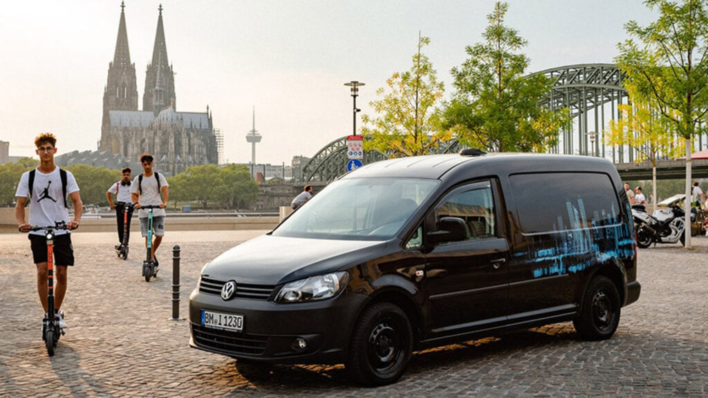Ein schwarzer INOVISCO Van steht am Rheinufer mit Blick auf den Kölner Dom und die Hohenzollernbrücke. Daneben fahren junge Leute auf E-Scootern vorbei. Die Szene verbindet urbane Mobilität mit aufmerksamkeitsstarker Werbung.