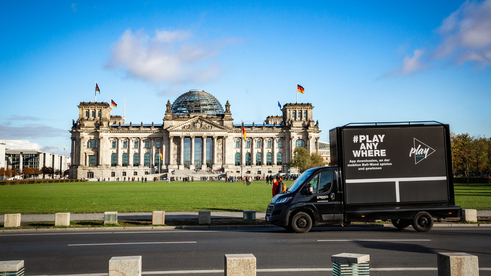 Ein schwarzer Werbetruck mit der Aufschrift „#PLAY ANYWHERE“ steht vor dem Reichstagsgebäude in Berlin. Im Hintergrund sind die Reichstagskuppel, deutsche Flaggen und eine große Grünfläche mit Besuchergruppen zu sehen.