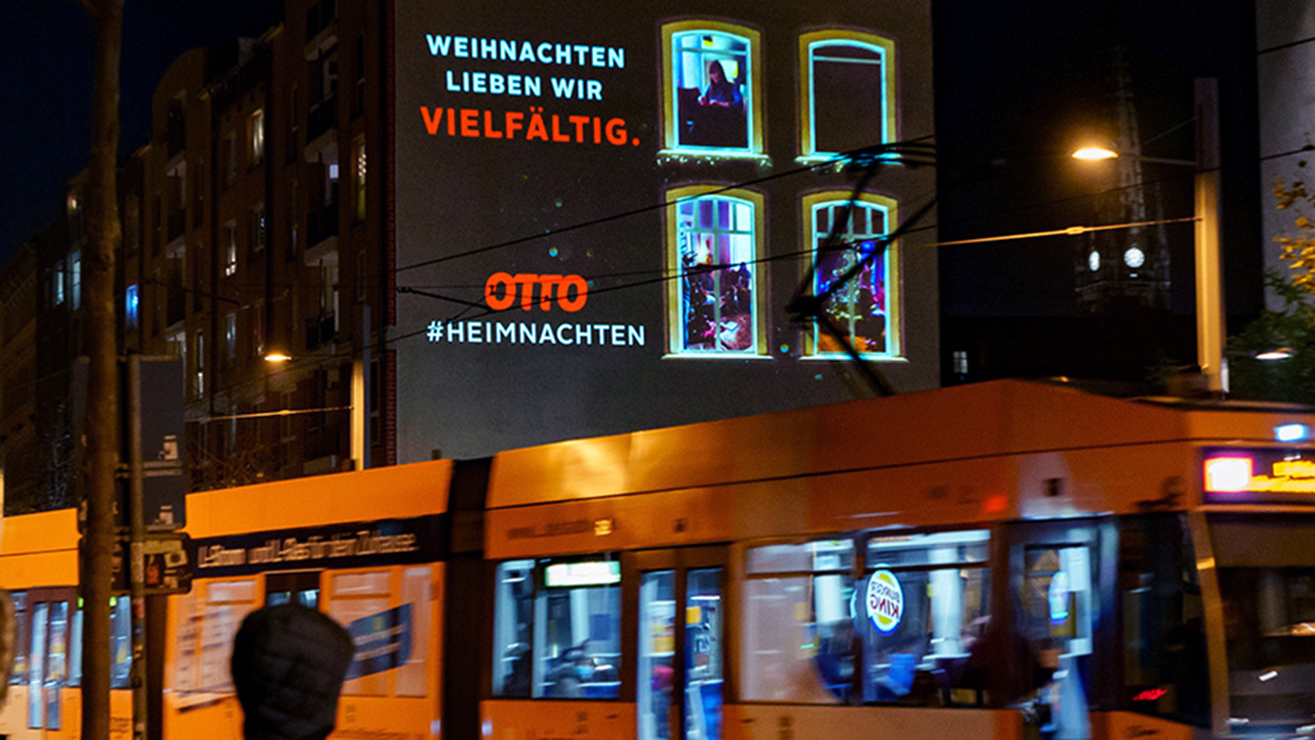 Eine großflächiges Digitales Riesenposter zeigt eine weihnachtliche Kampagne von OTTO mit dem Slogan „Weihnachten lieben wir vielfältig“. Leuchtende Fensterbilder verstärken die Botschaft. Im Vordergrund fährt eine Straßenbahn durch die nächtliche Stadt.