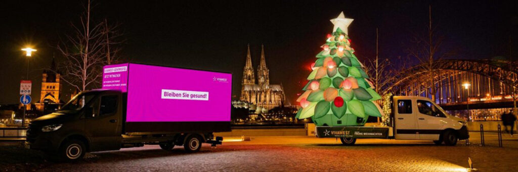 Das Bild zeigt einen INOVISCO DigiTruck bei Nacht am Rheinufer in Köln. Links steht der schwarze DigiTruck mit einer großen, pinken LED-Fläche, auf der der Text „Bleiben Sie gesund!“ zu lesen ist. Daneben befindet sich ein weiteres Fahrzeug mit einem großen, beleuchteten Weihnachtsbaum samt Stern und Kugeln, das ebenfalls die Aufschrift „VIVAWEST – Frohe Weihnachten“ trägt. Im Hintergrund sind der Kölner Dom und die Hohenzollernbrücke beleuchtet sichtbar.