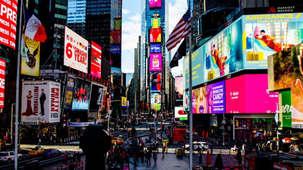 Eine belebte Szene am Times Square in New York mit unzähligen bunten Leuchtreklamen und riesigen digitalen Werbetafeln. Menschenmengen bewegen sich zwischen den Straßen, während Autos und Taxis durch die Kreuzungen fahren. Die grellen Displays prägen das Bild der Hochhäuser ringsum.