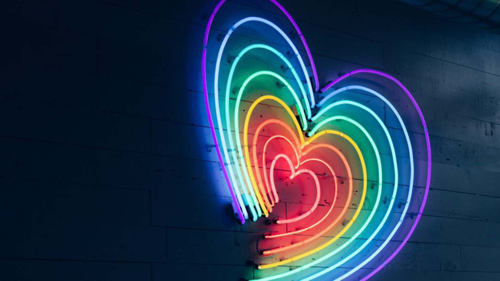 Das Bild zeigt ein leuchtendes Neonschild in Herzform, das aus mehreren übereinanderliegenden Herzlinien besteht. Jede Linie strahlt in einer anderen Farbe des Regenbogenspektrums – von Rot über Orange, Gelb, Grün, Blau bis Violett. Das Schild hängt an einer dunklen Wand, wodurch die kräftigen Neonfarben besonders intensiv hervorstechen.