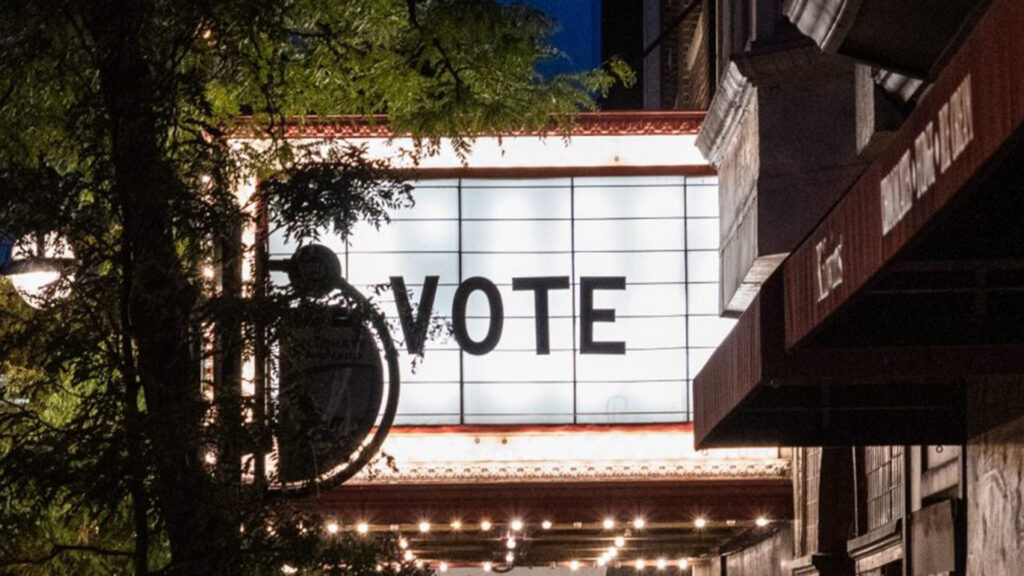 Ein beleuchtetes Kinomarquee trägt in großen schwarzen Buchstaben die Botschaft „VOTE“. Umrahmt von Lichterketten und Bäumen setzt das Schild ein klares Statement im nächtlichen Straßenbild.