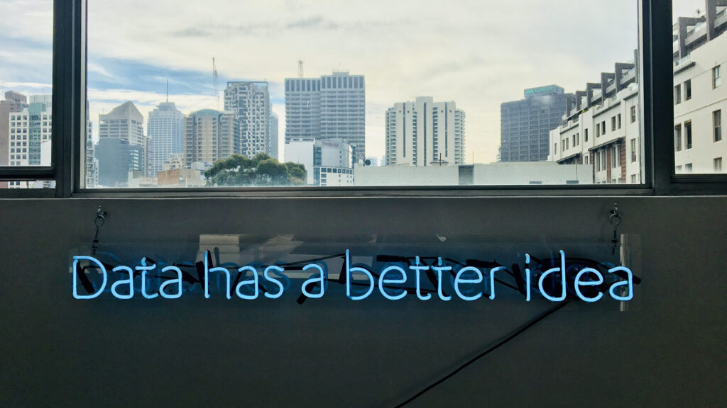 Ein leuchtendes Neon-Schild zeigt den Schriftzug „Data has a better idea“. Dahinter öffnet sich der Blick durch große Fenster auf eine Skyline mit modernen Hochhäusern.