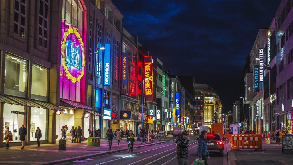 Eine belebte Einkaufsstraße am Abend mit leuchtenden Fassadenwerbungen und bunten Schildern bekannter Marken. Menschen spazieren und fahren mit dem Rad durch die Fußgängerzone. Die bunte Lichtkulisse sorgt für eine lebendige Innenstadt-Atmosphäre.