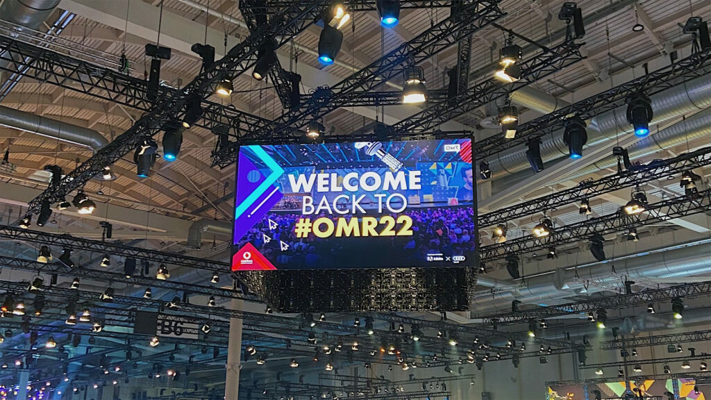 Ein großer Screen hängt unter der Hallendecke der OMR-Messe und zeigt die Begrüßung „Welcome back to #OMR22“. Im Hintergrund sind Scheinwerfer und Bühnentechnik zu sehen. Die LED-Fläche sorgt für maximale Aufmerksamkeit im Messeambiente.