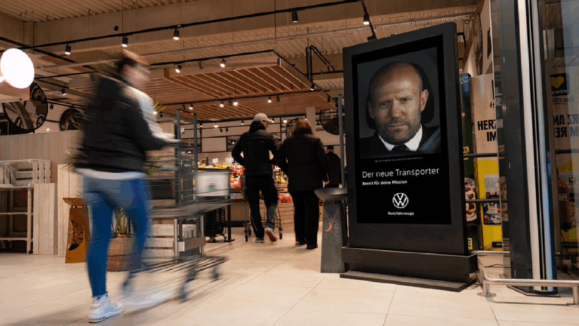Ein digitales Citylight Poster (DCLP) im Eingangsbereich eines Supermarkts zeigt eine VW-Werbung mit dem Slogan „Der neue Transporter – Bereit für deine Mission“. Einkaufende mit Wagen laufen an dem großen INOVISCO Werbemedium vorbei. Im Hintergrund sind Obst- und Gemüseabteilung sowie weitere Kunden erkennbar.