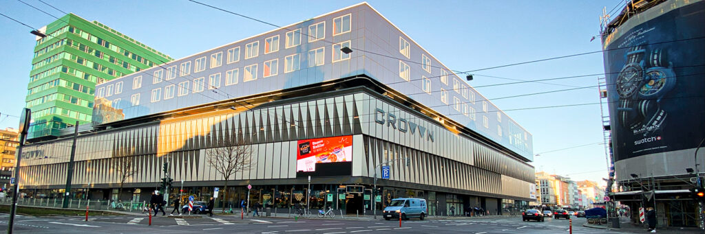 Auf dem Bild ist ein großflächiges DOOH-Display (Digital Out-of-Home) an der Fassade des Crown-Gebäudes in einer Innenstadtlage zu sehen. Auf dem Screen läuft gerade eine Lebensmittel-Werbung (Pizza-Angebot). Links im Bild fällt das auffällige grüne Hochhaus ins Auge, rechts ein riesiges Plakat für Swatch/Blancpain Uhren. Vor dem Gebäude kreuzen Straßenbahnschienen und eine vielbefahrene Kreuzung, was den Standort zu einem hochfrequentierten Werbepunkt macht. Ein Beispiel für aufmerksamkeitsstarke digitale Außenwerbung im urbanen Raum.