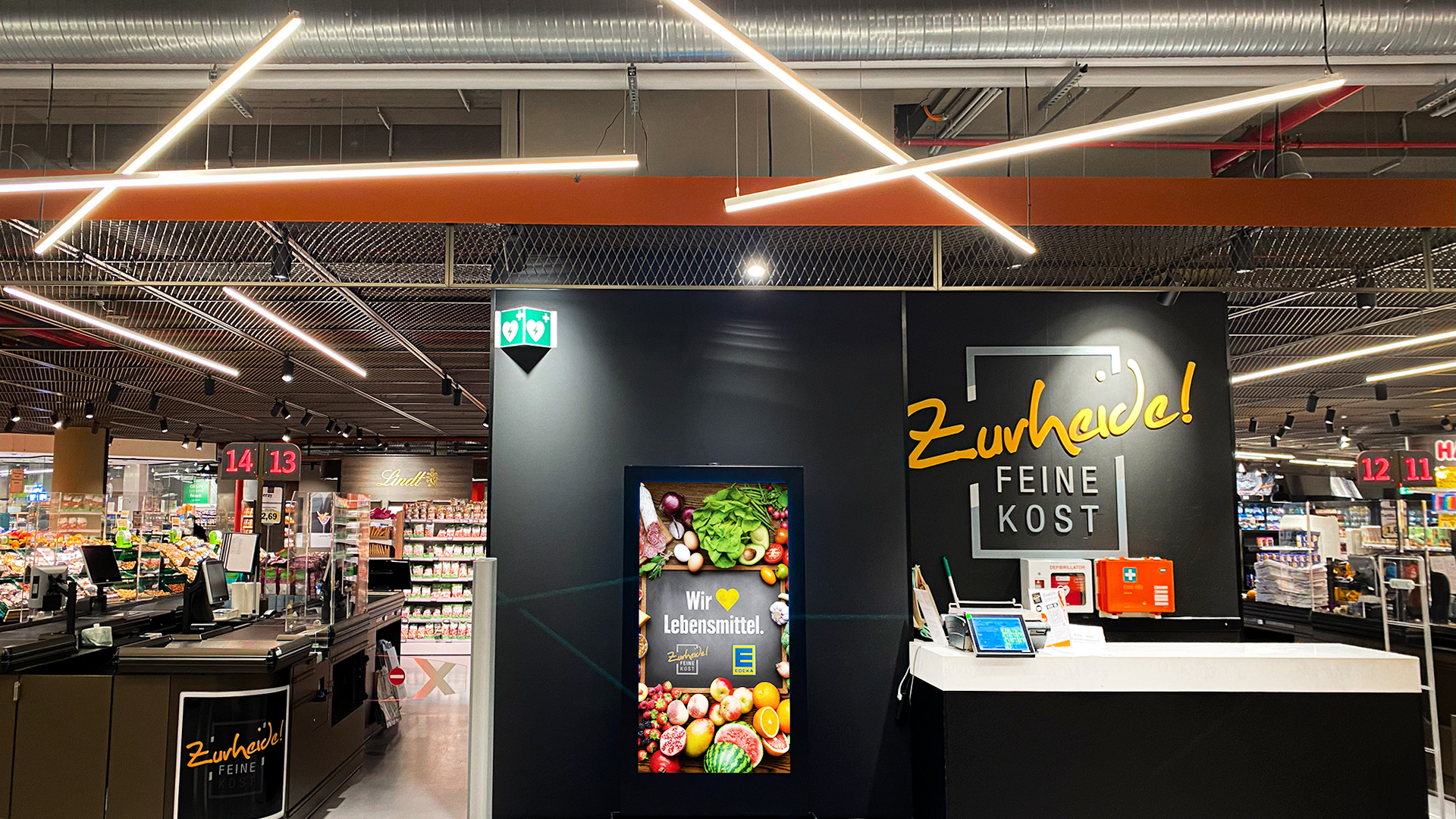 Ein digitales Citylight Poster (DCLP) im Eingangsbereich von Zurheide Feine Kost zeigt frisches Obst und Gemüse mit dem Slogan „Wir lieben Lebensmittel“. Daneben sind die modernen Kassenbereiche und Regalreihen des Marktes zu erkennen.