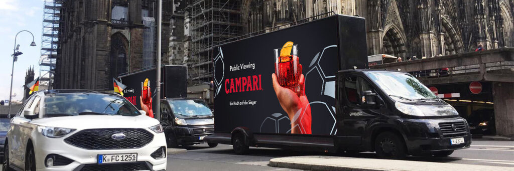 Ein schwarzer CoolLiteTruck von INOVISCO fährt durch die Innenstadt und zeigt eine große Campari-Werbung mit einem roten Drinkglas und der Aufschrift „Public Viewing – Ein Hoch auf die Sieger“. Im Hintergrund ragt der Kölner Dom empor, während Autos und Passanten die Szene beleben.