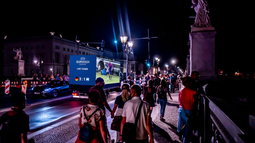 Ein DigiTruck von INOVISCO fährt nachts durch eine belebte Innenstadt und zeigt live den Spielstand der UEFA EURO 2024. Auf dem großen Bildschirm ist das Ergebnis Deutschland gegen Schottland mit 3:1 eingeblendet. Zahlreiche Menschen laufen über die Straße und verfolgen das Geschehen in lebendiger Atmosphäre.