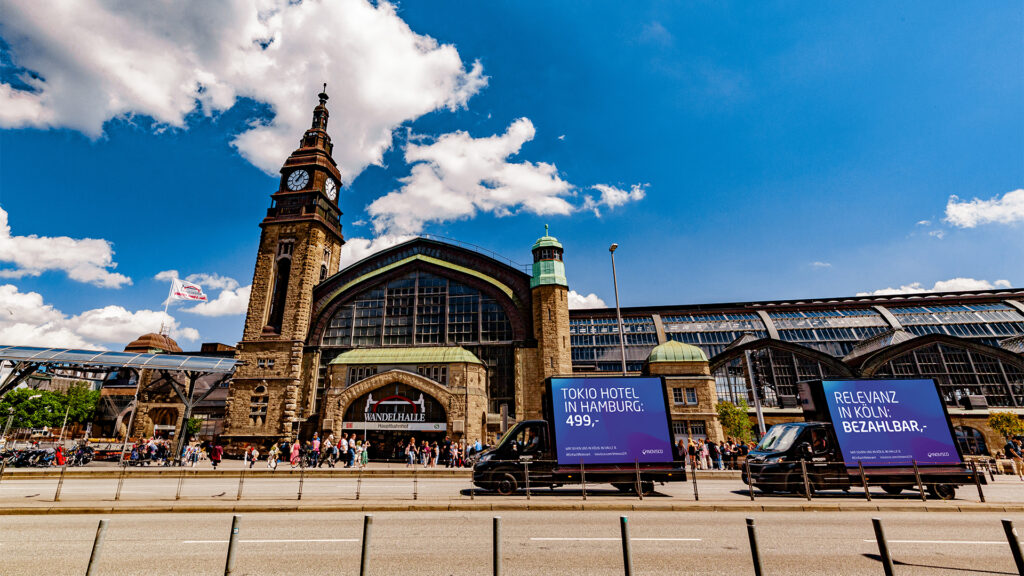 Vor dem Hamburger Hauptbahnhof stehen zwei INOVISCO CoolLiteTruck mit auffälligen, blauen Werbeflächen. Die mobilen Plakate sind direkt an der belebten Straße positioniert und werden von zahlreichen Passant:innen und vorbeifahrendem Verkehr gesehen.