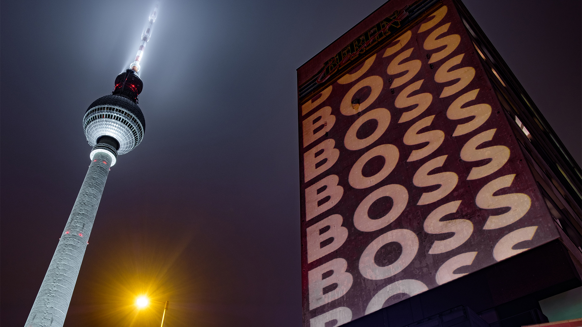 Der Berliner Fernsehturm leuchtet in den Nachthimmel, daneben ist die Fassade eines Gebäudes großflächig mit dem Schriftzug „BOSS“ in Wiederholung angestrahlt. Eine Straßenlaterne setzt zusätzlich gelbes Licht in die Szene.