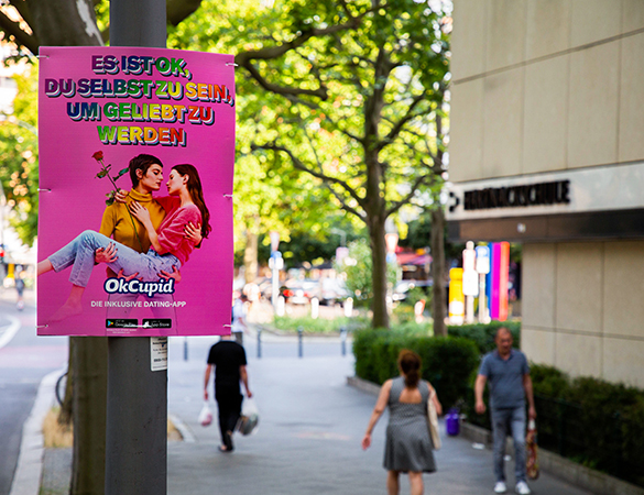 Ein buntes INOVISCO Hohlkammerplakat der Dating-App OkCupid hängt an einer Straßenlaterne. Der Slogan „Es ist ok, du selbst zu sein, um geliebt zu werden“ steht über einem Motiv mit zwei sich küssenden Personen. Fußgänger passieren die belebte Szene in der Innenstadt.