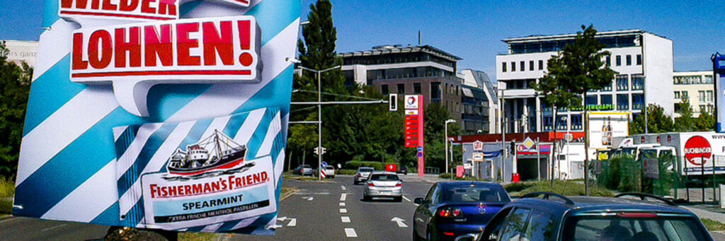 Ein INOVISCO Hohlkammerplakat für Fisherman’s Friend ist an einem Laternenpfahl an einer vielbefahrenen Straße angebracht. Vorbeifahrende Autos und umliegende Gebäude unterstreichen die starke Sichtbarkeit der Werbefläche.
