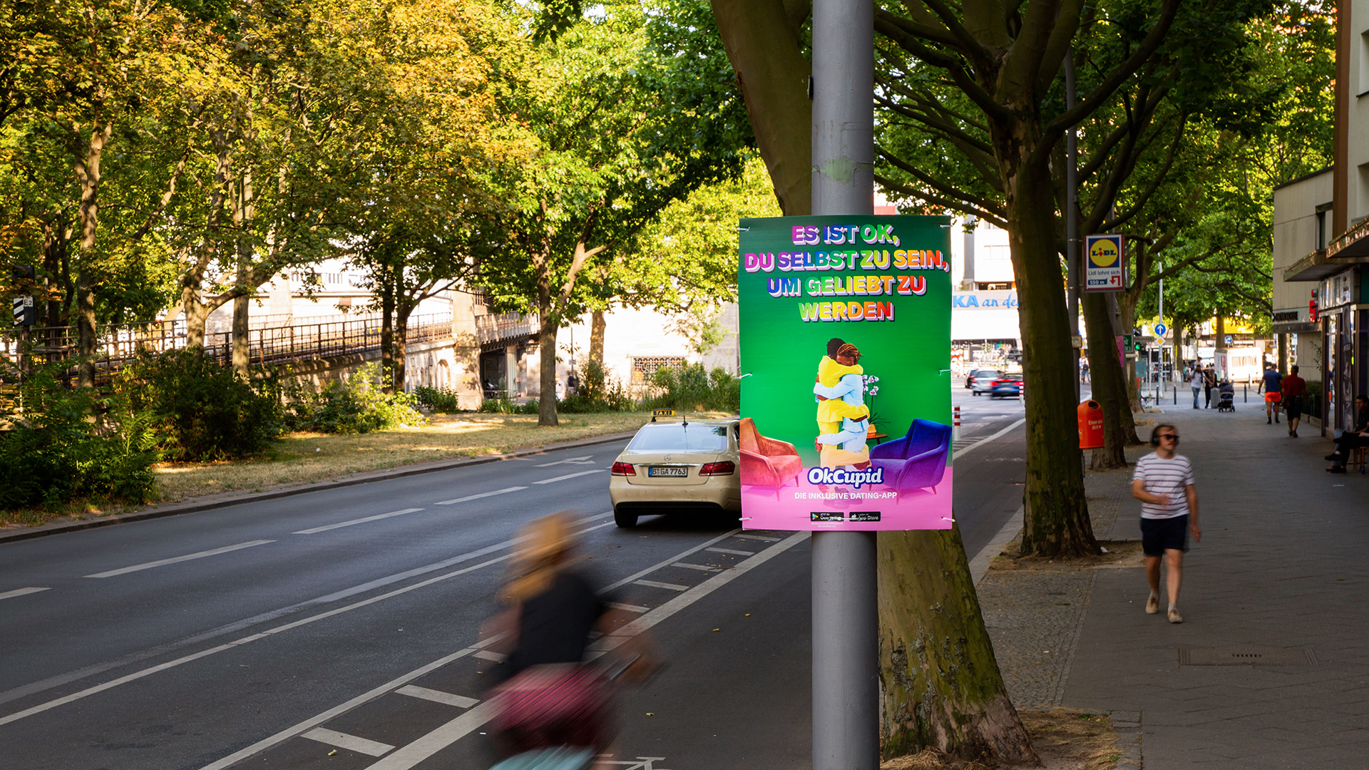 Ein farbenfrohes INOVISCO Hohlkammerplakat von OkCupid hängt an einer Straßenlaterne auf einer belebten Allee. Darauf umarmen sich zwei Personen mit dem Slogan „Es ist ok, du selbst zu sein, um geliebt zu werden“. Fußgänger und Radfahrer passieren die Szene im Sommerlicht.