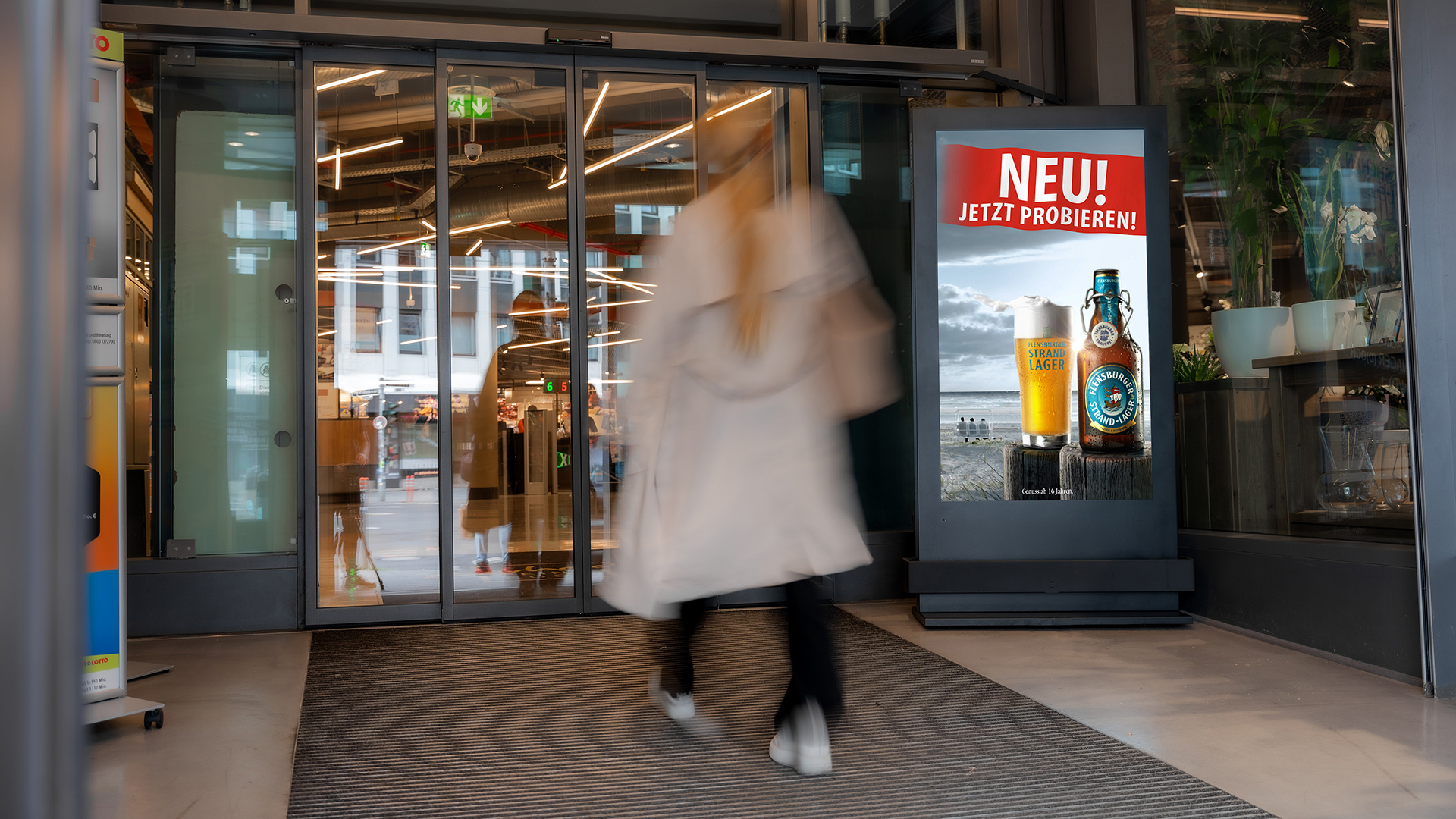 Ein INOVISCO DCLP im Eingangsbereich eines Supermarkts zeigt eine Flensburger Strand Lager Werbung mit dem Schriftzug „NEU! Jetzt probieren!“. Kundinnen und Kunden betreten das Geschäft und passieren dabei die digitale Werbefläche.