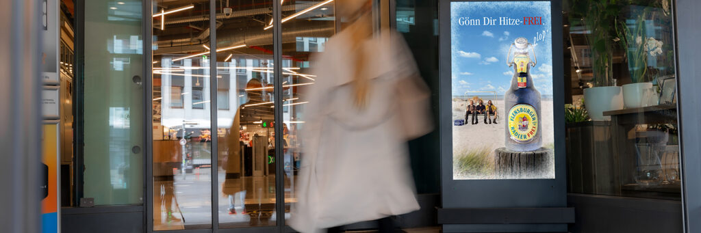 Ein INOVISCO DCLP im Eingangsbereich eines Supermarkts zeigt eine Flensburger-Werbung mit der Botschaft „Gönn dir Hitze-FREI“. Vorbeigehende Kundinnen und Kunden passieren die digitale Werbefläche beim Betreten des Geschäfts.