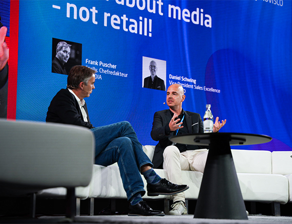 Zwei Männer sitzen auf einer hellen Bühne auf weißen Sesseln und sprechen miteinander. Im Hintergrund ist eine große blaue Leinwand mit Text und Porträtfotos zu sehen. Auf dem Tisch zwischen ihnen steht eine Wasserflasche.
