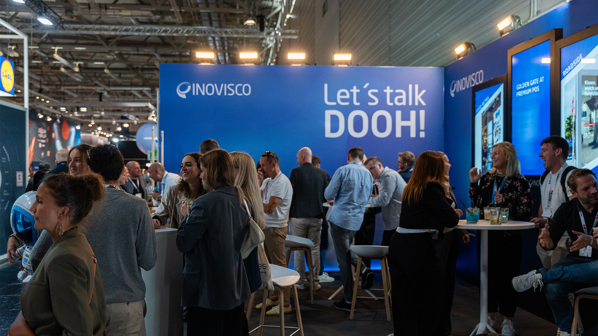 Menschen stehen und unterhalten sich an einem Messestand mit blauen Wänden. Groß an der Rückwand prangt der Schriftzug „Let’s talk DOOH!“ neben dem INOVISCO-Logo. Rechts sind große Bildschirme mit weiteren Informationen eingebaut, während Besucher Getränke in der Hand halten.
