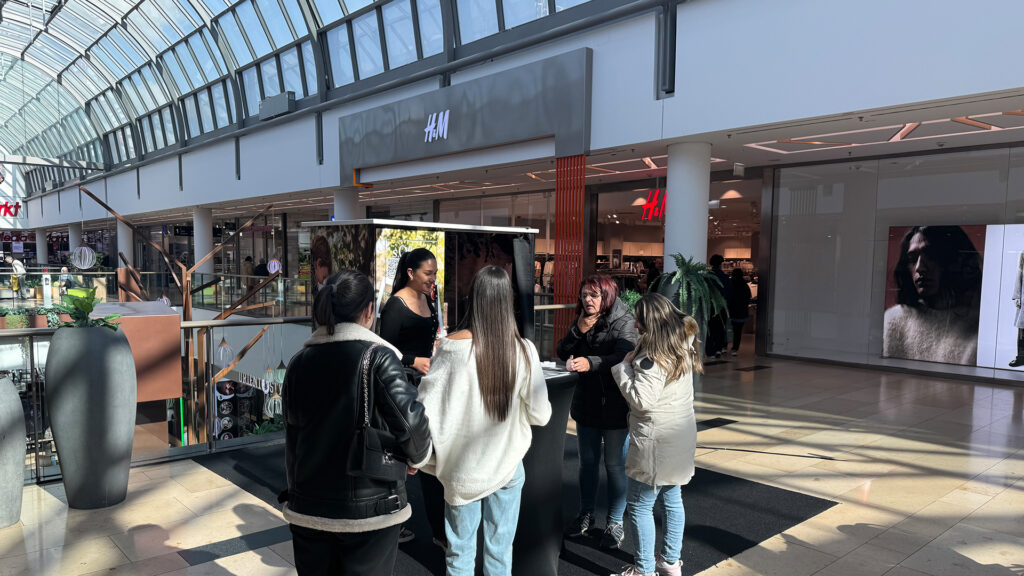 Ein Promotionstand von s.Oliver ist im Eingangsbereich eines Einkaufszentrums aufgebaut. Eine Promoterin spricht mit interessierten Besucherinnen, die sich um den Stand versammelt haben. Im Hintergrund sind H&M-Filiale und weitere Geschäfte zu sehen.