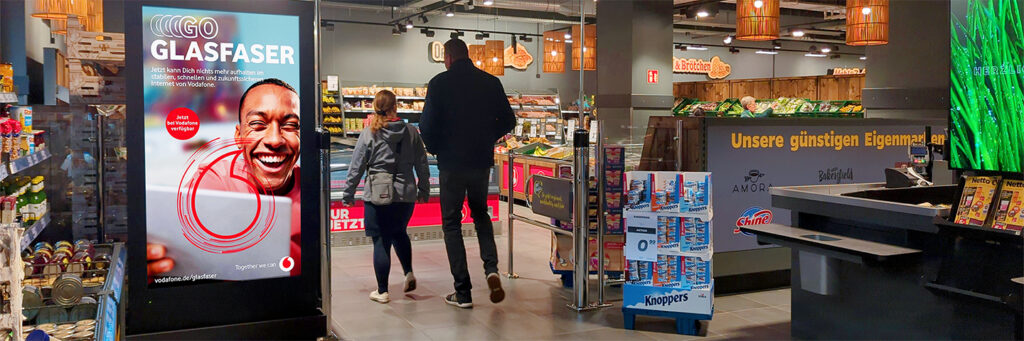 Ein INOVISCO DCLP im Eingangsbereich eines Supermarkts zeigt eine Vodafone-Werbung für Glasfaser-Internet. Kunden mit Einkaufswagen passieren den digitalen Werbeträger auf dem Weg in die Gemüse- und Backwarenabteilung.