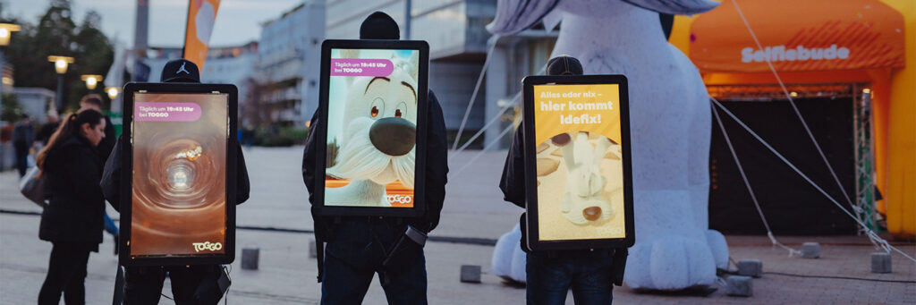 Drei Personen tragen DigiBoards mit digitalen Werbeflächen für TOGGO. Die Motive zeigen unter anderem den Hund Idefix und bunte Animationen. Im Hintergrund steht eine große aufblasbare Figur mit der Aufschrift „Selfiebude“.