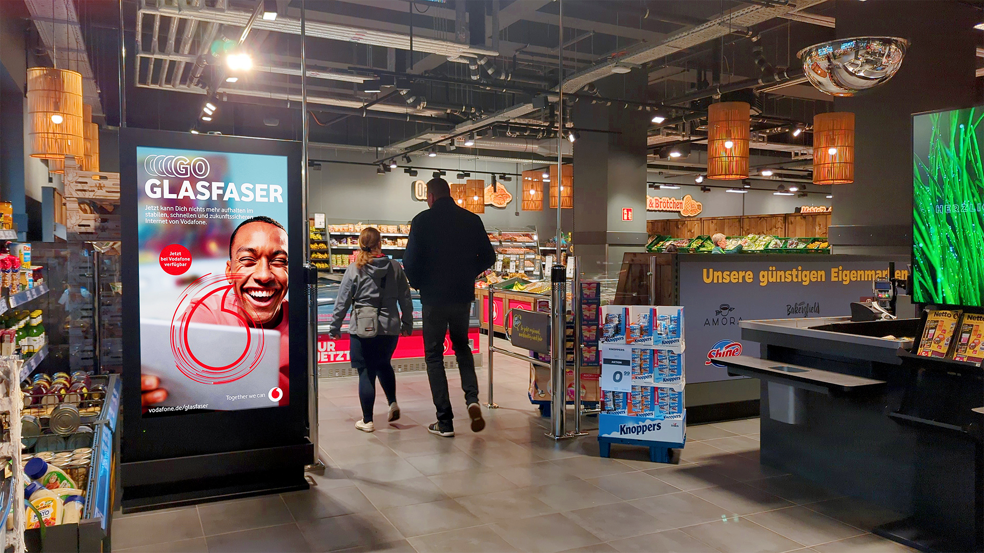Ein INOVISCO DCLP im Eingangsbereich eines Supermarkts zeigt eine auffällige Vodafone-Werbung für Glasfaser. Kunden betreten den Markt und passieren den digitalen Werbeträger auf dem Weg zu den Regalen.