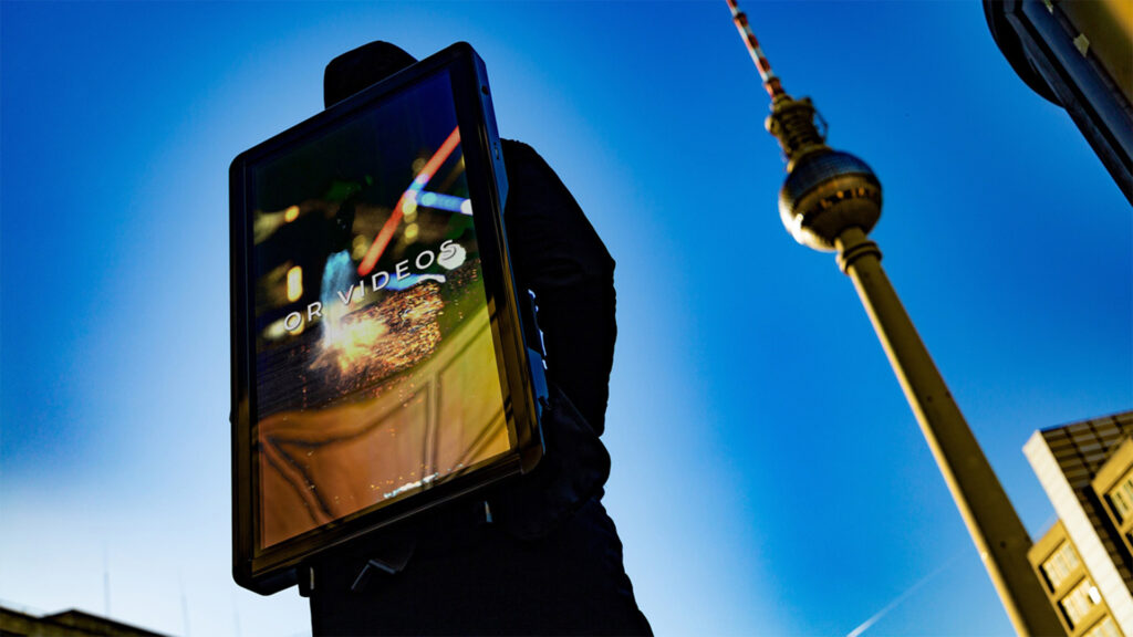 Eine Person trägt ein leuchtendes DigiBoard mit Videomotiv vor dem Berliner Fernsehturm. Die mobile DOOH-Werbefläche von INOVISCO fällt durch ihre Größe und Helligkeit auf. Klare Himmel und Stadtkulisse rahmen die Szene.
