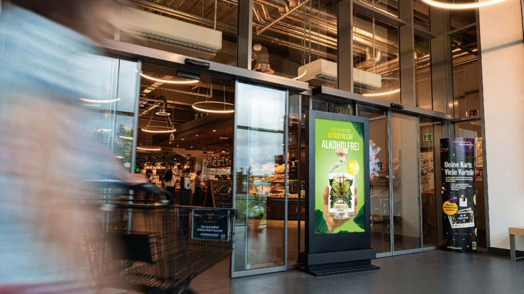 Ein INOVISCO DCLP am Eingang eines Supermarkts zeigt eine auffällige Anzeige für Siegfried Wonderleaf alkoholfrei. Kunden mit Einkaufswagen passieren den digitalen Werbeträger auf dem Weg in den Markt.