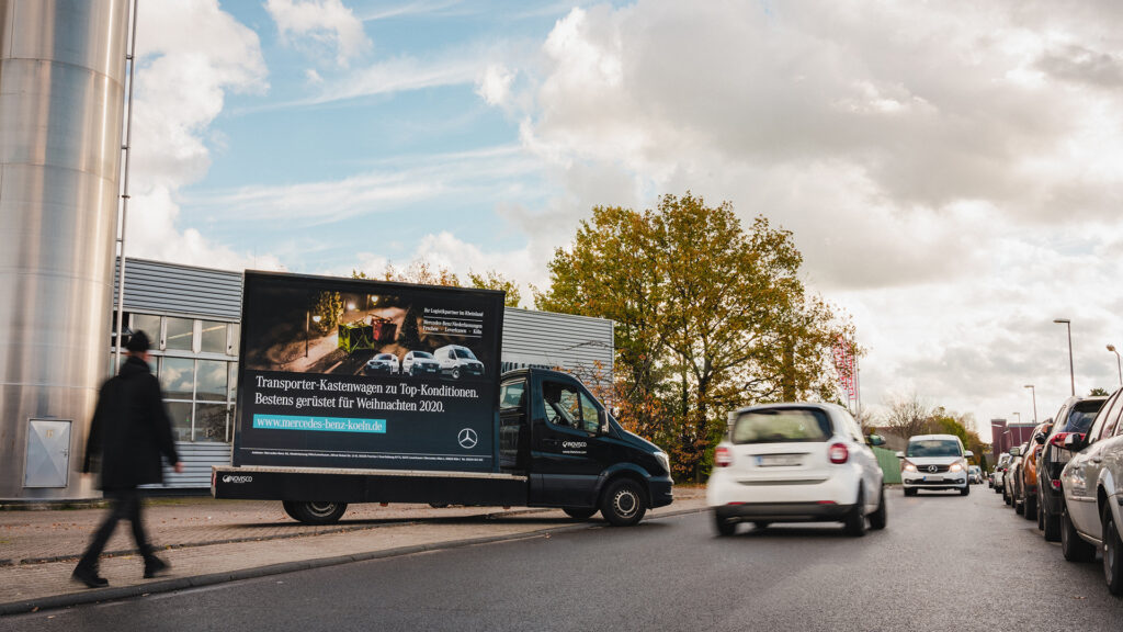 Ein INOVISCO LitoTube mit Mercedes-Benz Werbung steht am Straßenrand vor einem Autohaus. Das Plakat wirbt für Transporter-Kastenwagen zu Top-Konditionen und weihnachtlicher Bildsprache. Vorbeifahrende Autos und Passanten verstärken die Präsenz der mobilen Werbefläche.