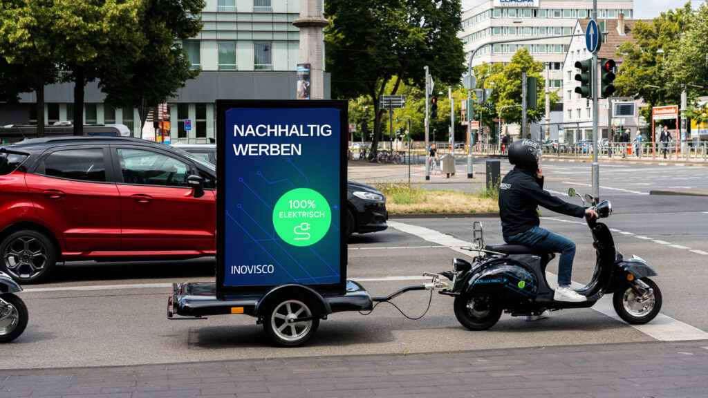 Ein INOVISCO CoolLiteScooter mit Anhänger wirbt mit der Aufschrift „Nachhaltig werben – 100% elektrisch“. An einer städtischen Kreuzung fährt der E-Scooter neben Autos und setzt ein Zeichen für umweltfreundliche Außenwerbung.