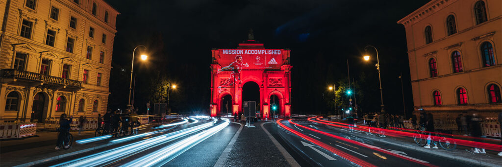 Ein historisches Stadttor ist nachts in kräftigem Rot angestrahlt und zeigt eine Projektion mit der Aufschrift „Mission Accomplished“ sowie einem Fußballspieler und dem Adidas-Logo. Vor dem Bauwerk ziehen die Lichter vorbeifahrender Autos lange Lichtspuren durch die Straße. Links und rechts rahmen beleuchtete Gebäude die Szene ein.