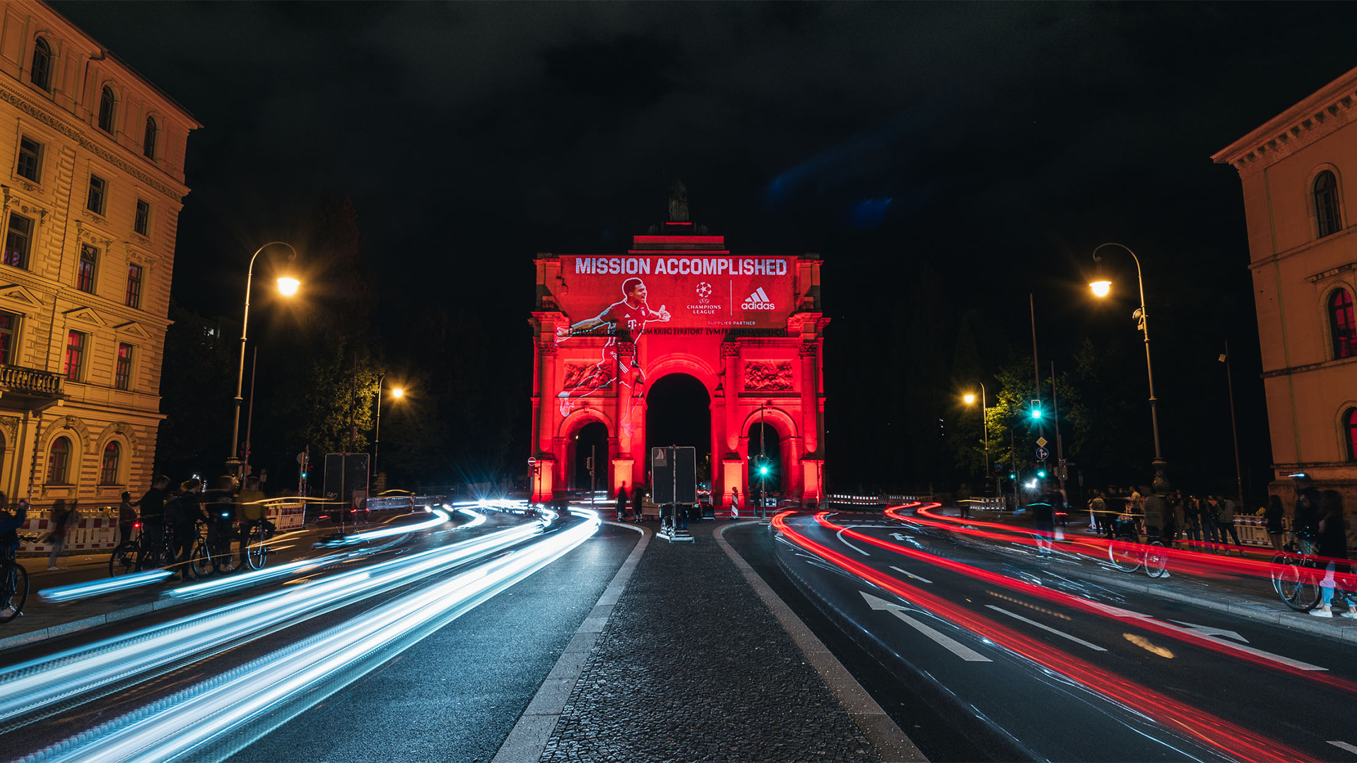Ein nachts rot angestrahltes Stadttor zeigt eine Projektion mit einem Fußballspieler, dem Schriftzug „Mission Accomplished“ und Logos von Champions League, Adidas und FC Bayern. Vor dem Bauwerk ziehen die Scheinwerfer vorbeifahrender Autos helle Lichtspuren durch die Straße. Links und rechts stehen historische Gebäude mit beleuchteten Fenstern.