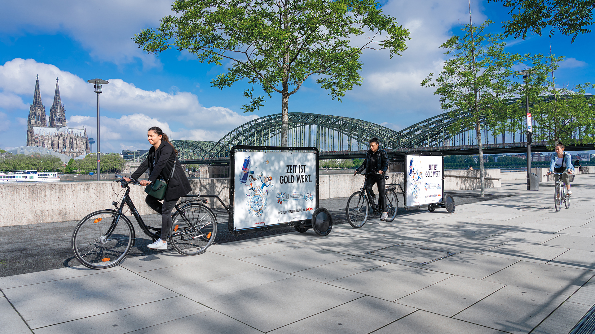 Zwei INOVISCO CoolLiteBikes rollen am Kölner Rheinufer entlang und tragen Red Bull Plakate mit dem Slogan „Zeit ist Gold wert“. Im Hintergrund erheben sich der Kölner Dom und die Hohenzollernbrücke. Die mobile Werbung fügt sich in die urbane Kulisse ein.