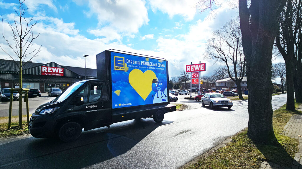 Ein CoolLiteTruck mit großflächiger EDEKA-Werbung fährt vorbei – im Hintergrund ist deutlich ein REWE-Markt zu sehen.