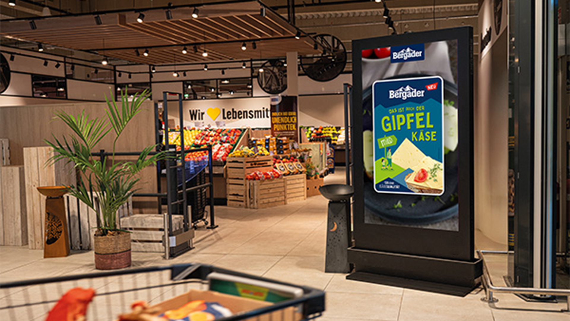 Ein digitales Citylight Poster (DCLP) im Eingangsbereich eines Supermarktes wirbt für Bergader Gipfelkäse. Im Vordergrund ist der Rand eines Einkaufswagens erkennbar, dahinter sind Obst- und Gemüsestände sowie ein Schild mit dem Slogan „Wir ♥ Lebensmittel“.