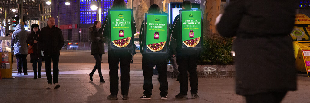 Drei Personen tragen DigiBoards von INOVISCO in einer Innenstadt bei Nacht. Die leuchtenden Werbeflächen zeigen eine Kampagne von Kühne mit humorvollen Sprüchen und Pizzamotiven. Passanten bewegen sich rundherum durch die Straße.