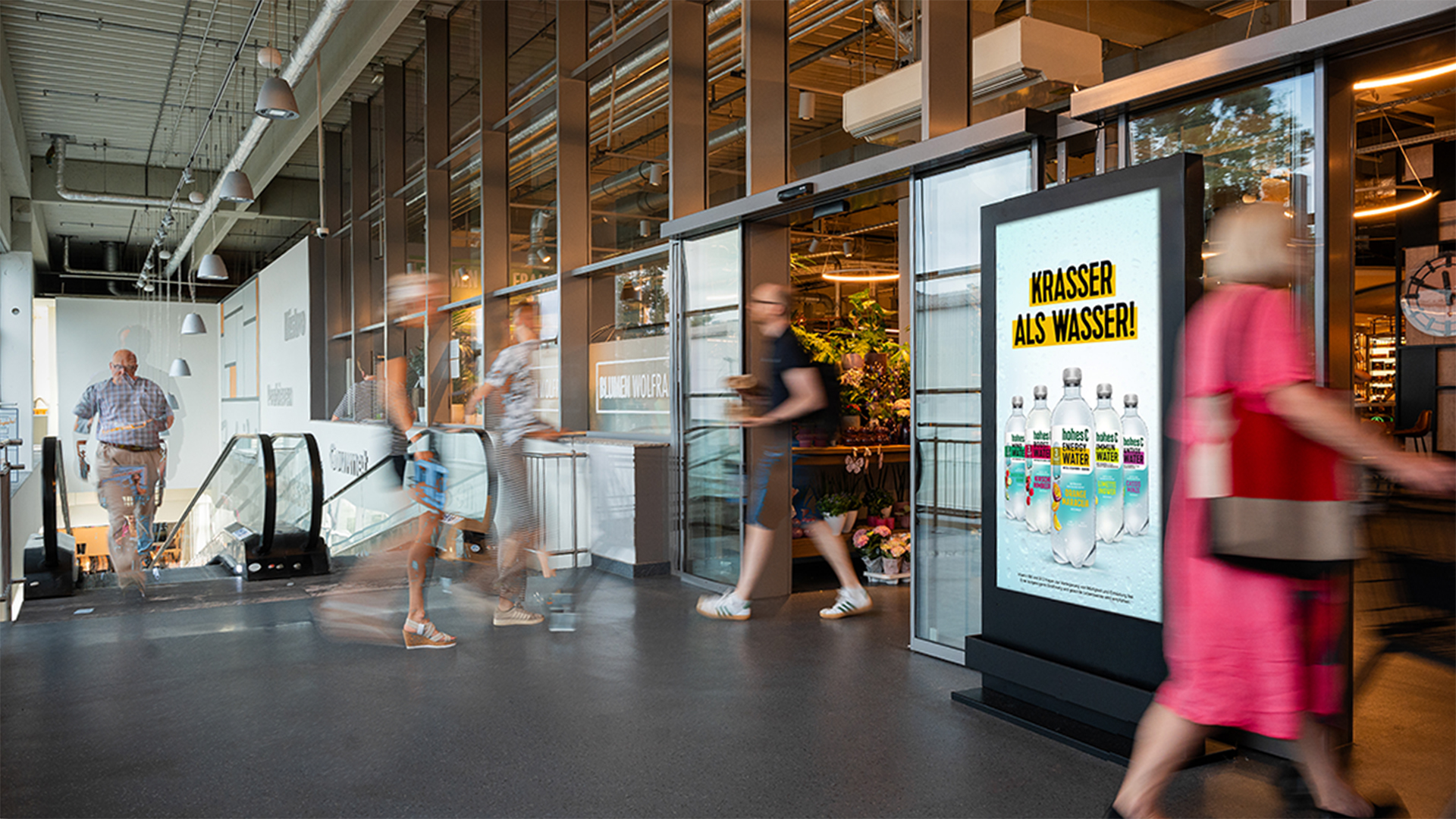 Das Bild zeigt ein DCLP-Display direkt am Eingang eines Supermarktes. Auf dem Screen läuft eine Honest Energy Water Werbung mit dem auffälligen Slogan „Krasser als Wasser!“. Passant:innen bewegen sich dynamisch ins und aus dem Geschäft, was die Lebendigkeit und Frequenz des Ortes unterstreicht.